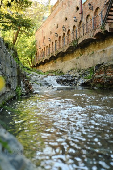 A clear stream flows calmly past old brick structures surrounded by lush greenery. Sunlight