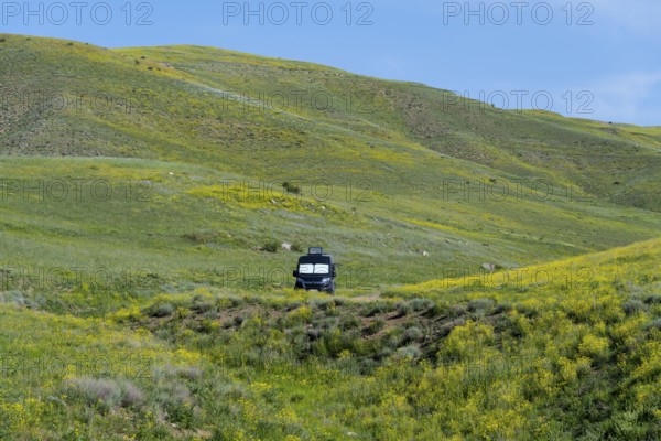 A black van stands alone in a meadow next to green and yellow hills under a blue sky, camper,