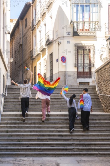 Young adults are joyfully walking up stone stairs in a european city, carrying rainbow flags and a