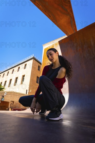 Young woman squatting on an urban surface, posing casually against a modern rust colored