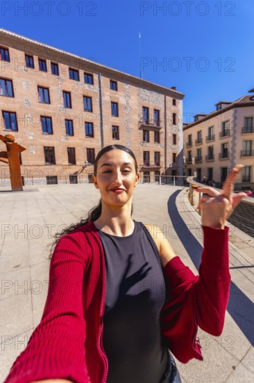 Young woman with dark hair posing for a selfie in a city square, holding her hand up, with old