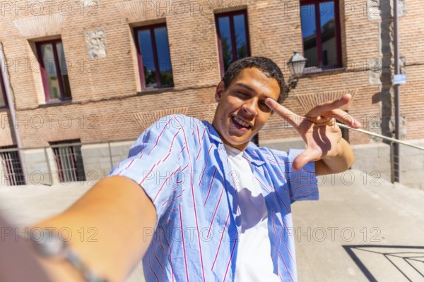 Young man smiling and making a playful hand gesture while taking a selfie in sunny urban streets,