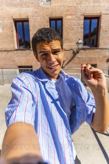 Young man smiling directly at the camera while taking a selfie outdoors on a sunny day, wearing a