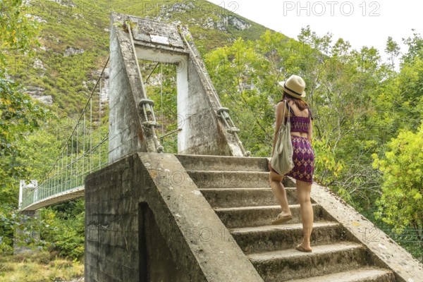 Female tourist walking up concrete stairs leading to an old suspension bridge over the dobra river