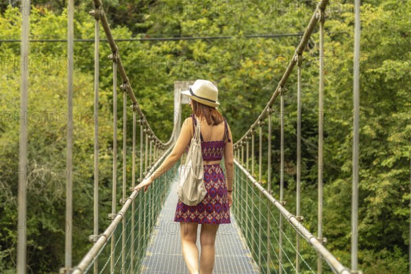 Female tourist with hat and backpack walking on a metal suspension bridge, enjoying the scenic