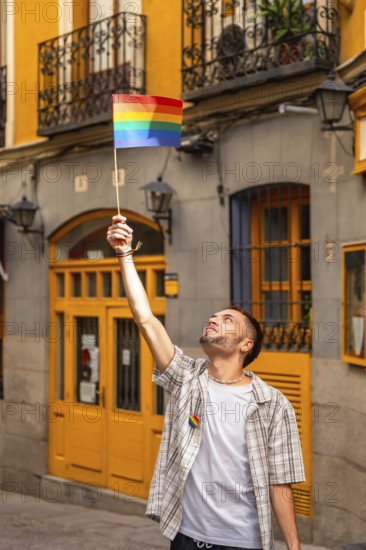 Young man standing in a city street, raising a small rainbow flag with arm outstretched,