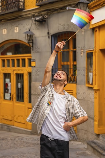 Young man standing on a street, smiling and raising a small rainbow flag, expressing happiness and