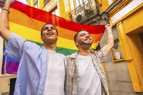 Happy young men joyfully embracing and holding a colorful rainbow flag together, looking up in a