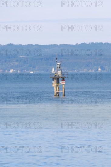 A lighthouse in blue water with a sailing ship, green hills and clear sky in the background,