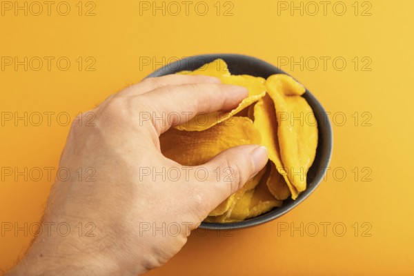 Dried Mango in blue ceramic bowl with hand on orange pastel paper background. Side view, close up.