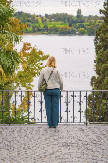 Person standing at the railing of a walkway and looking at a lake surrounded by trees in autumn,