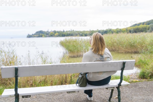 Woman on park bench looking at calm lake and autumn landscape, Mainau Island, Lake Constance,