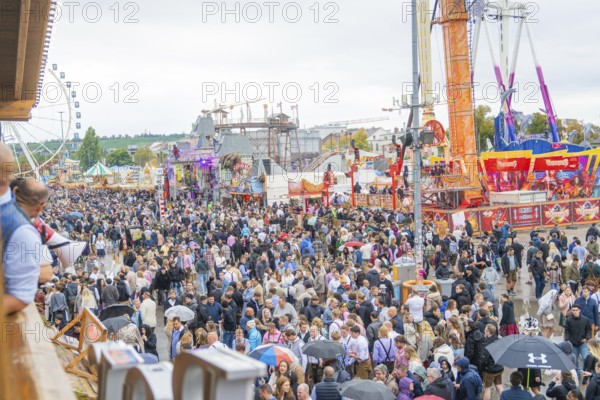Large crowd at a folk festival with fairground rides under a cloudy sky, Cannstatter Wasen folk