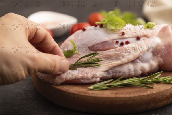 Raw Turkey Thigh with spices and rosemary on cutting board on black concrete background with hand