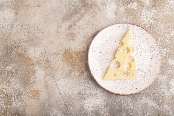 Piece of cheese with holes on ceramic plate on brown concrete background, top view, flat lay, copy