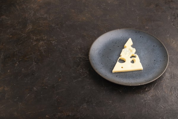 Piece of cheese with holes on blue plate on black concrete background, side view, copy space,