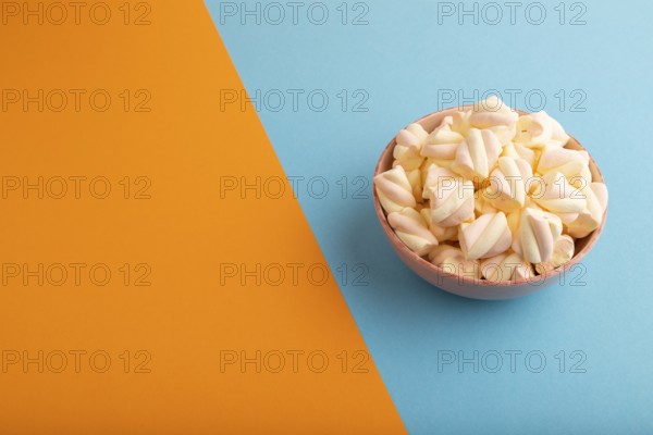 Orange and pink marshmallow in ceramic bowl on blue and orange pastel paper background, side view,