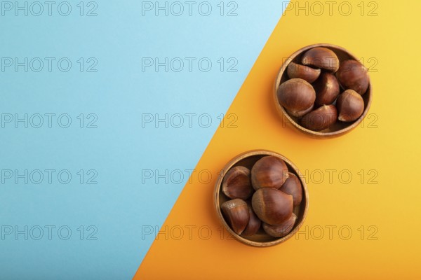 Wooden bowl with raw edible ?hestnuts on blue and orange paper pastel background, top view, flat