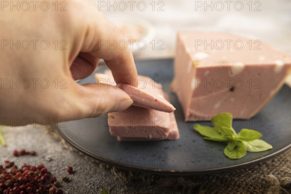 Mortadella sausage with hand on plate with pepper and herbs on brown concrete background and linen