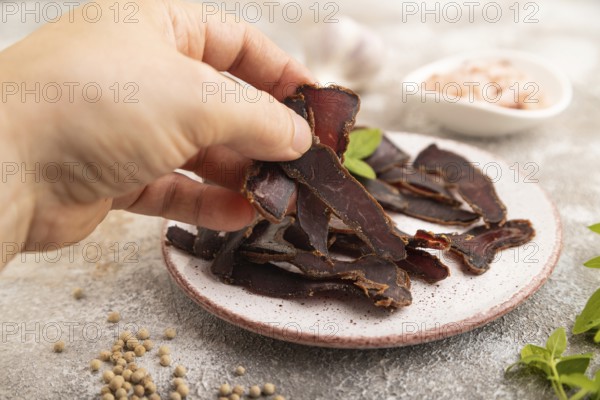 Armenian Basturma dried meat with hand on plate with pepper and herbs on brown concrete background.