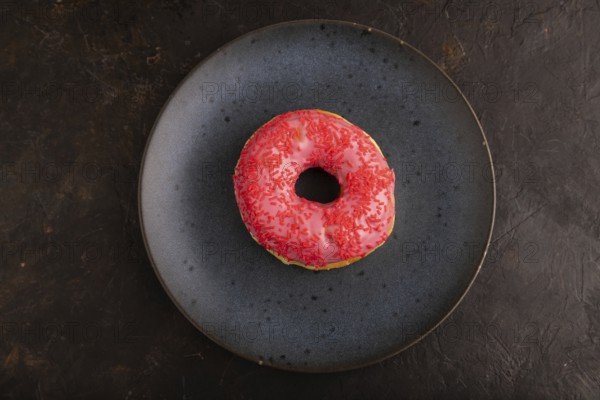 Pink Donut with sprinkles on blue ceramic plate on black concrete background, top view, flat lay,