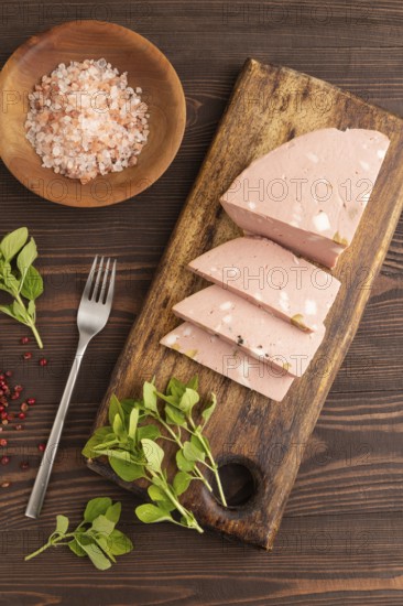 Mortadella sausage on wooden cutting board with pepper and herbs on brown wooden background. Top