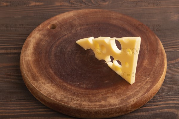 Piece of cheese with holes on cutting board on brown wooden background, side view, close up,