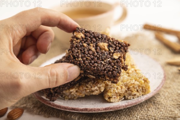 Granola bars with caramel, nuts, flakes in ceramic plate with hand on white wooden background,