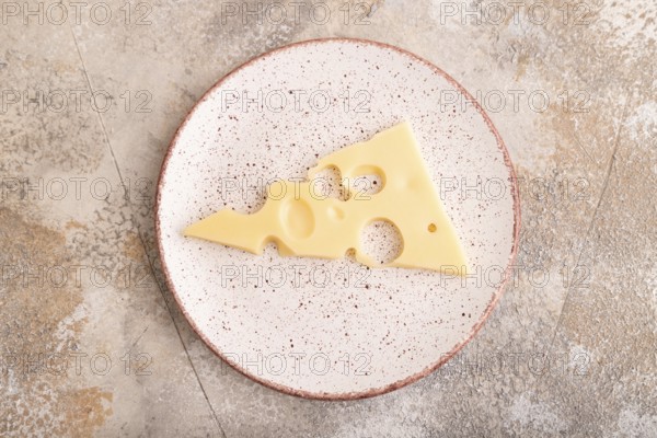 Piece of cheese with holes on ceramic plate on brown concrete background, top view, flat lay, copy
