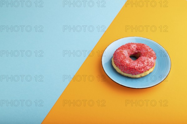 Pink Donut with sprinkles on blue ceramic plate on blue and orange pastel paper background, side