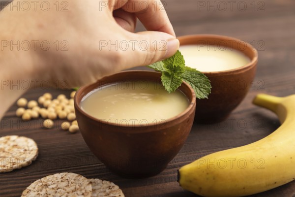 Organic non dairy banana and soy milk in clay cup with hand on brown wooden background. Vegan