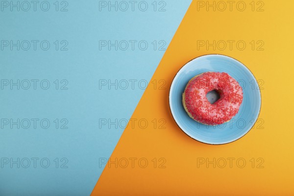 Pink Donut with sprinkles on blue ceramic plate on blue and orange pastel paper background, top