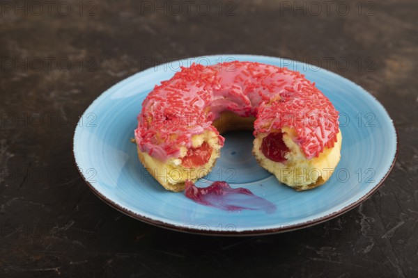 Bitten Pink Donut with sprinkles on blue ceramic plate on black concrete background, side view,