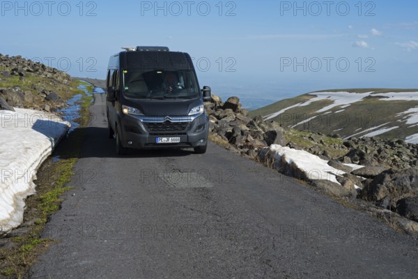 Van fährt auf asphaltiertem Weg neben Felsen und Schneefeldern mit klarem Himmel, das Tal im