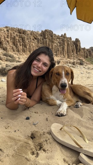 Young woman and her furry companion smiling, lying on sandy beach under an umbrella, enjoying a