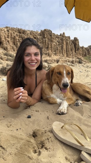 Young smiling woman with dark hair lying on a sandy beach next to a happy brown dog, both relaxing