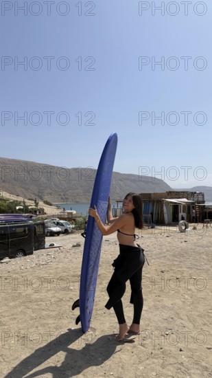 Young woman smiling, holding a blue surfboard on a dusty beach, wearing a wetsuit top and bottom,