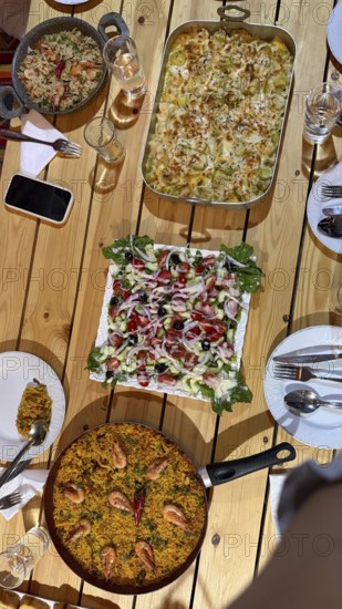 Rustic wooden table set with multiple homemade dishes including a large pan of seafood paella, a