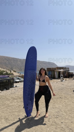 Young woman on a sunny moroccan beach holding a blue surfboard, wearing a bikini top and wetsuit