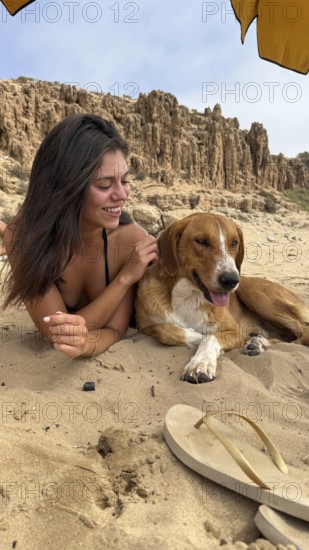 Woman lying on sand, smiling and gently petting a dog, relaxing on a beach with cliff formations in