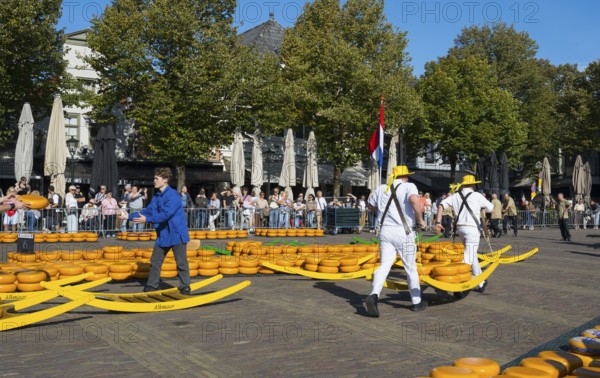 Männer in traditionellen Trägeranzügen auf einem Käsemarkt unter einer niederländischen Flagge,