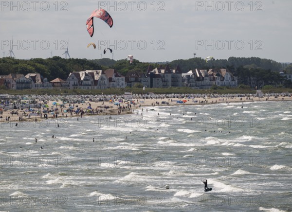 Kitesurfers and holidaymakers on the beach and in the sea, waves, beach chairs, houses in the