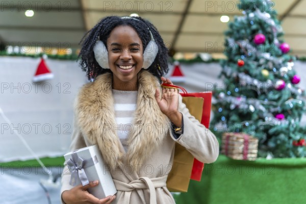 Young black woman wearing earmuffs holding christmas present and shopping bags, smiling in front of