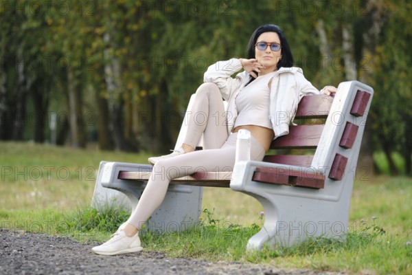 A woman sits on a park bench dressed in light-colored casual clothes, sunglasses on, with a relaxed