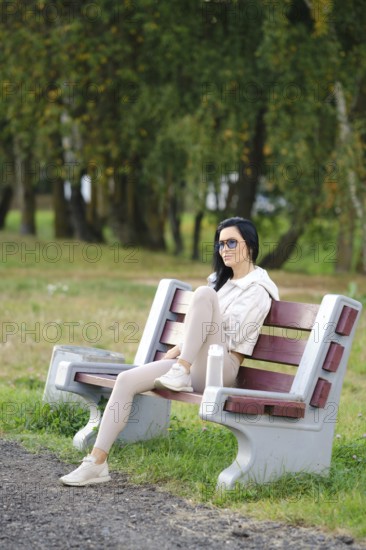 A slender woman sits comfortably on a park bench, wearing glasses and casual attire. Sunlight