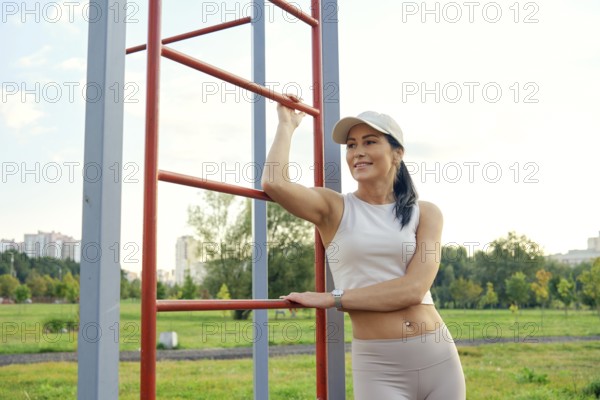 A fitness enthusiast relaxes at a park, leaning against outdoor exercise equipment. She enjoys the