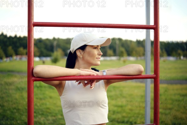 A woman in a casual outfit stands at a fitness station in a park, resting her arms on the bars