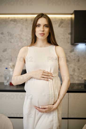 A pregnant woman stands in a contemporary kitchen, gently cradling her baby bump. She has long hair