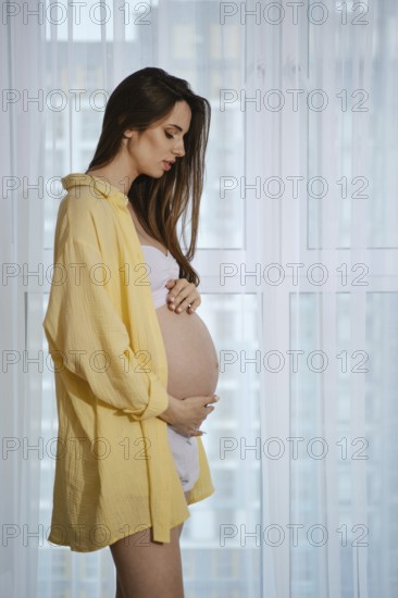 A pregnant woman stands near a window in profile, wearing a yellow shirt and white outfit, lovingly