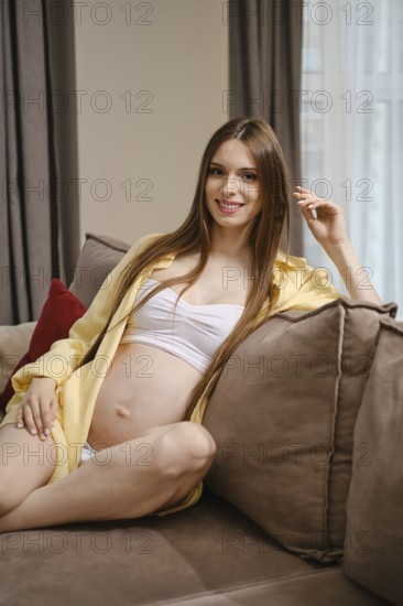 A relaxed expectant mother sits comfortably on a soft sofa, wearing a light outfit that highlights